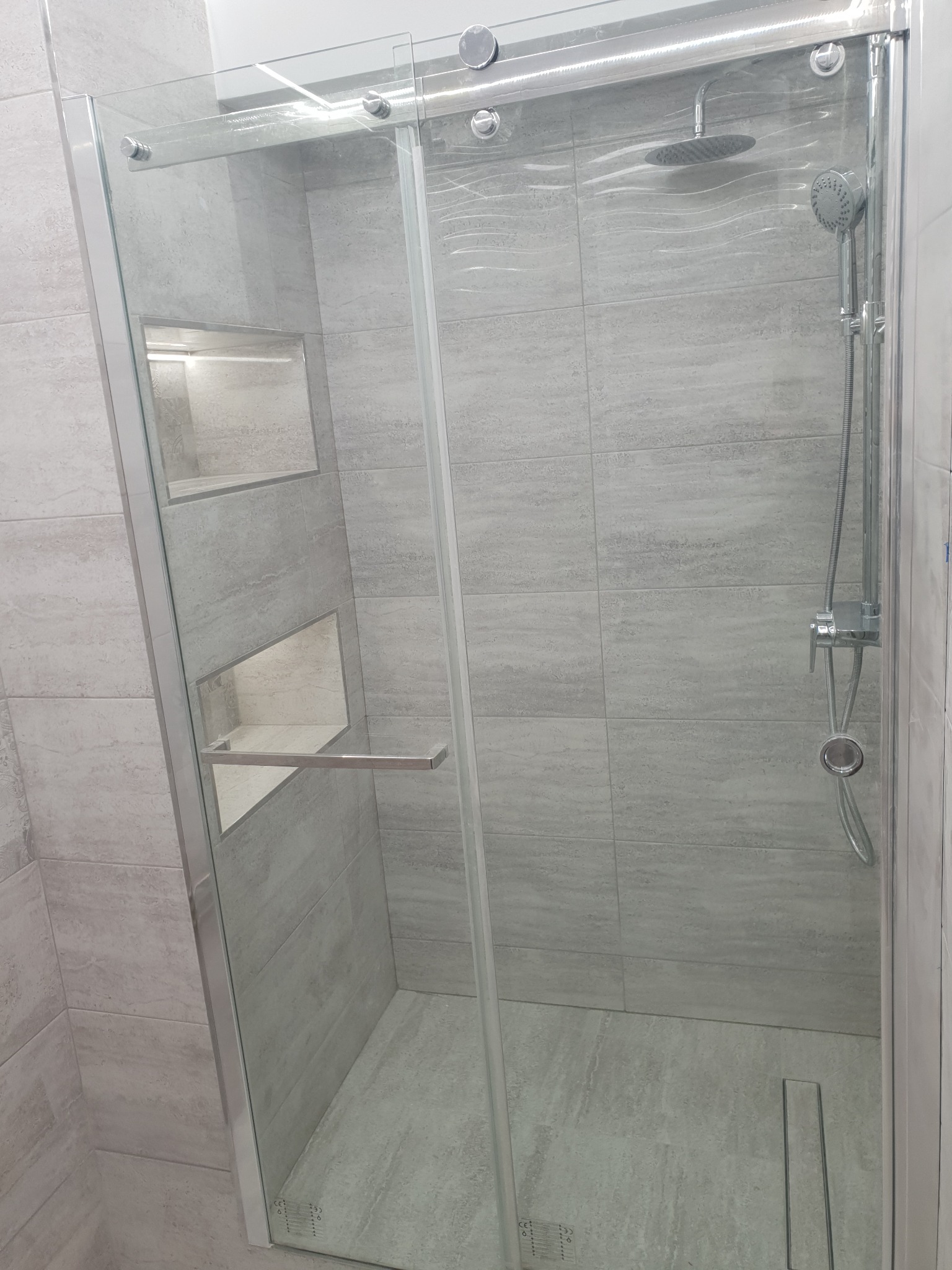 Nowoczesna kabina prysznicowa z przesuwnymi szklanymi drzwiami, wykończona jasnoszarą płytką imitującą kamień, z wnękami na kosmetyki i deszczownicą.