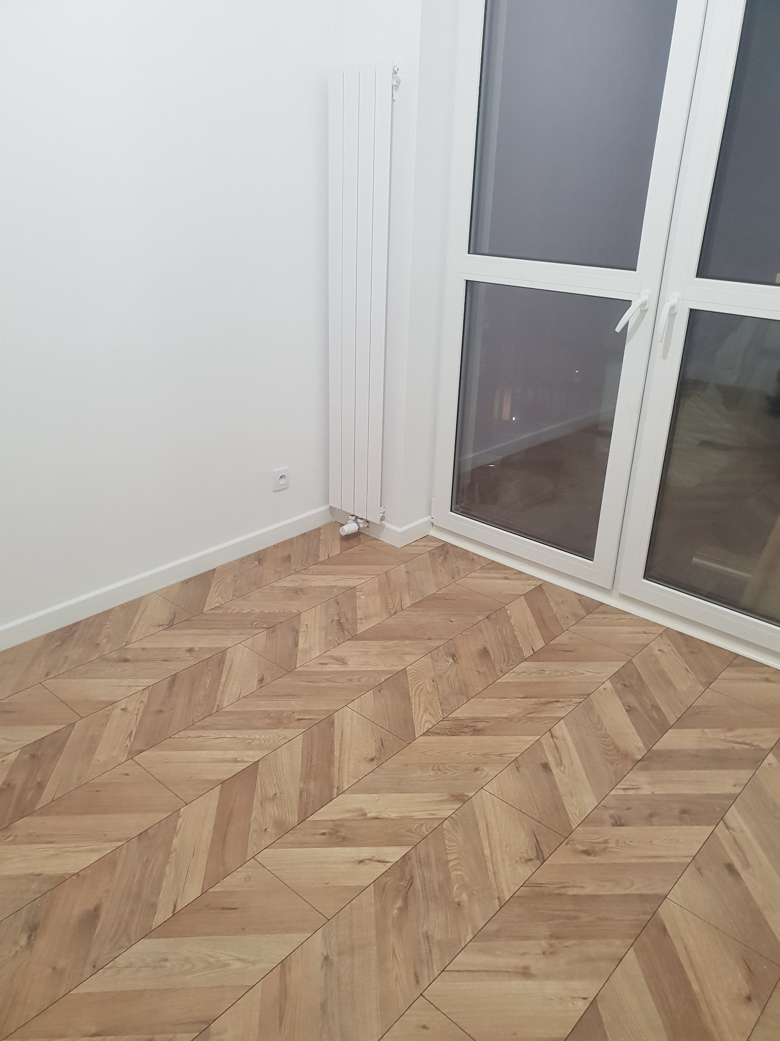 Ułożone panele podłogowe w jodełkę francuską, widok na białą ścianę z grzejnikiem i kontakt oraz białe okno z widokiem na zewnątrz.