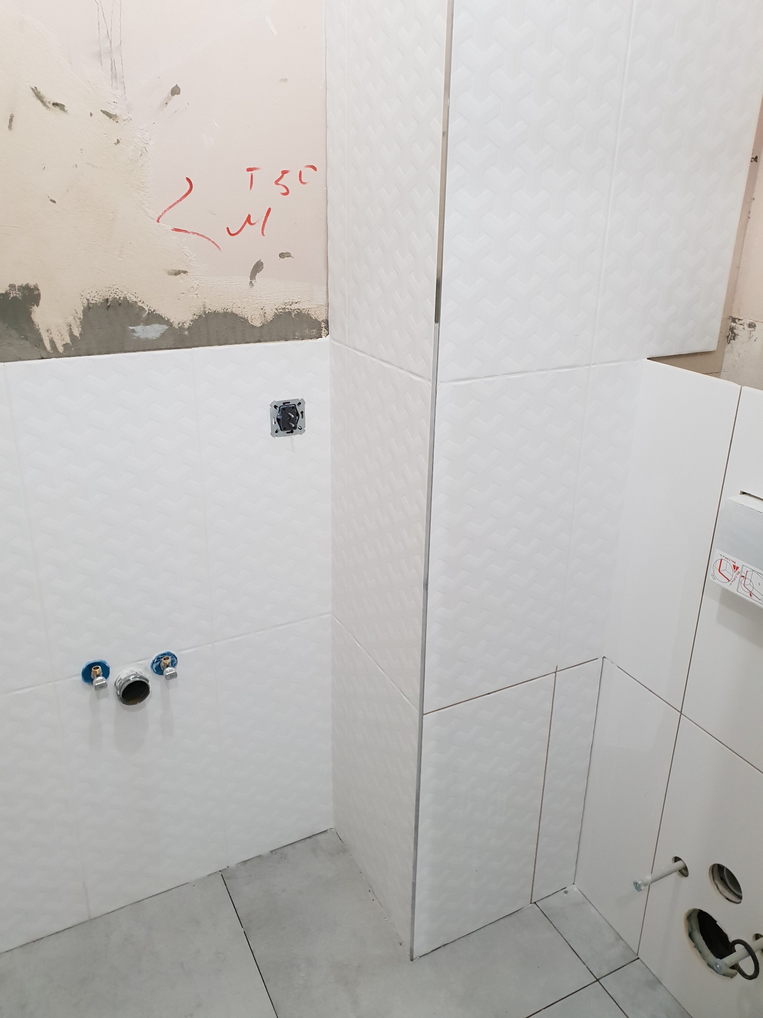 Wnętrze łazienki w trakcie prac wykończeniowych: białe płytki z delikatnym wzorem ułożone na ścianach, widoczne przyłącza wodne i otwór na kontakt, fragment ściany z resztkami starej farby...