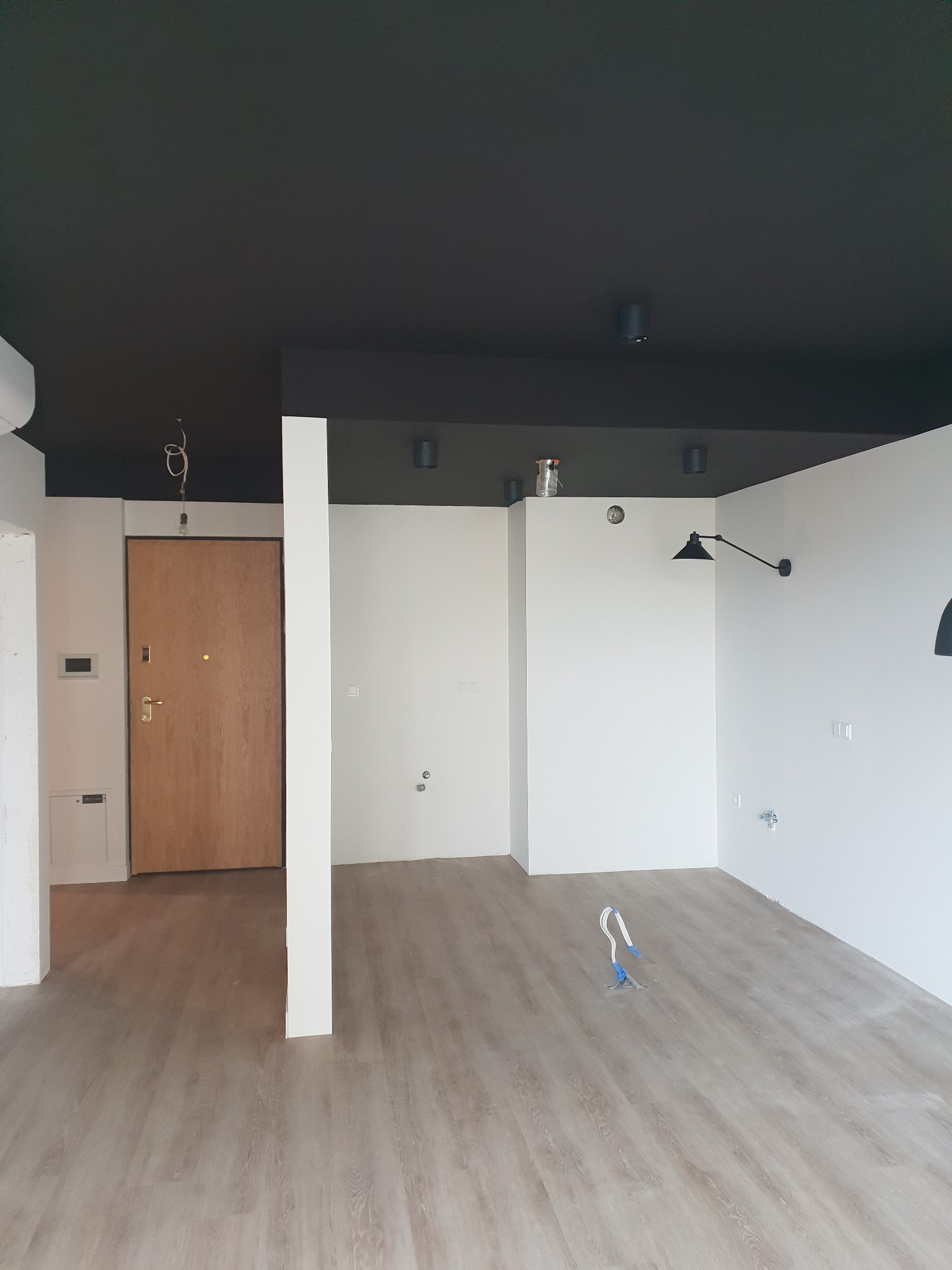 Wnętrze pokoju po remoncie z czarnym sufitem, jasnymi ścianami i panelami podłogowymi; widoczne elementy instalacji elektrycznej i niedokończone detale.