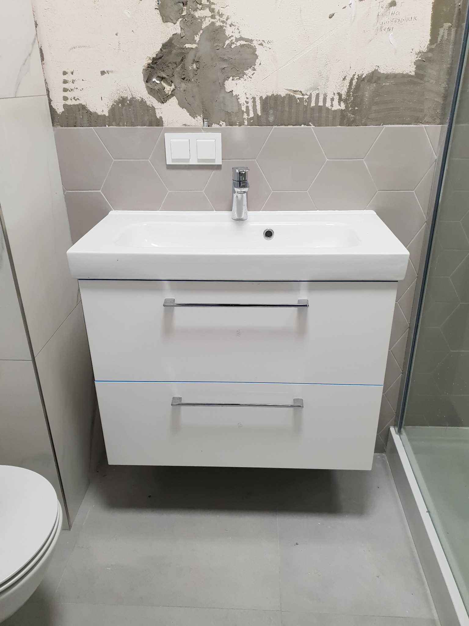 Biała szafka umywalkowa z chromowanymi uchwytami w łazience w trakcie remontu, widoczne szare heksagonalne płytki i fragmenty nieotynkowanej ściany.