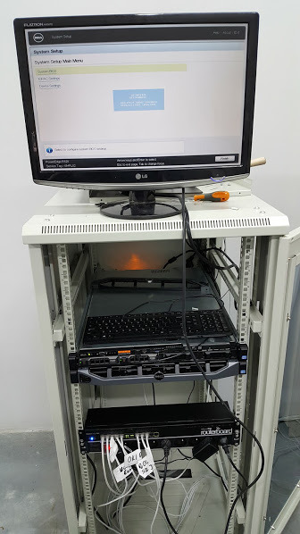 Serwerownia z monitorem wyświetlającym ekran konfiguracji systemu BIOS, klawiaturą i routerem z podłączonymi kablami oznaczonymi etykietami, umieszczona w szafie rack.