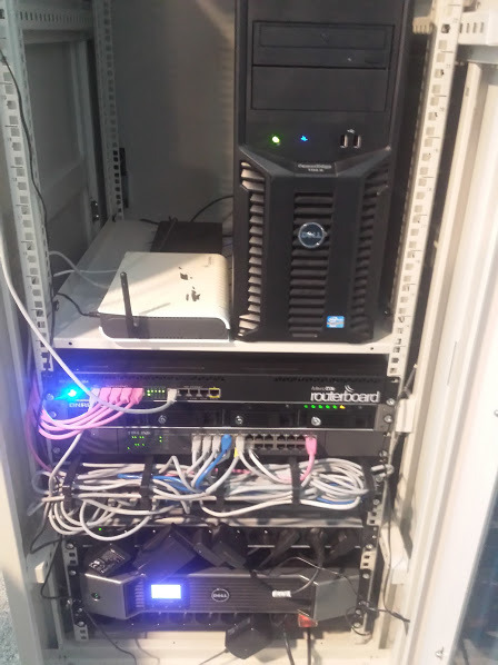 Szafa rack z serwerem Dell, routerem MikroTik Routerboard, switchem i punktem dostępowym Wi-Fi, okablowanie strukturalne z różowymi kablami Ethernet.