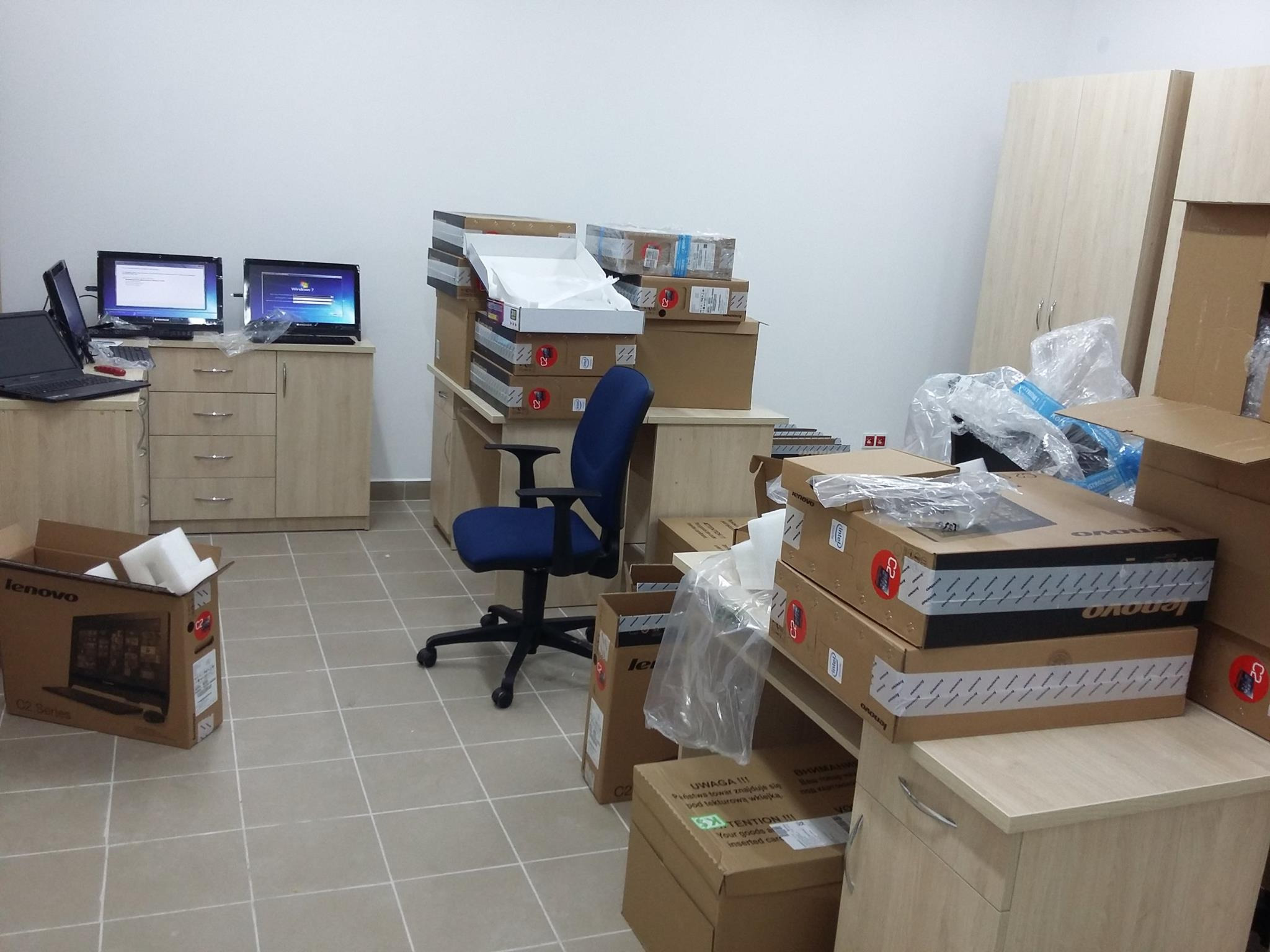 Biuro po dostawie nowego sprzętu komputerowego: monitory i laptopy Lenovo w kartonach, przygotowane do instalacji i konfiguracji, krzesło biurowe pośrodku