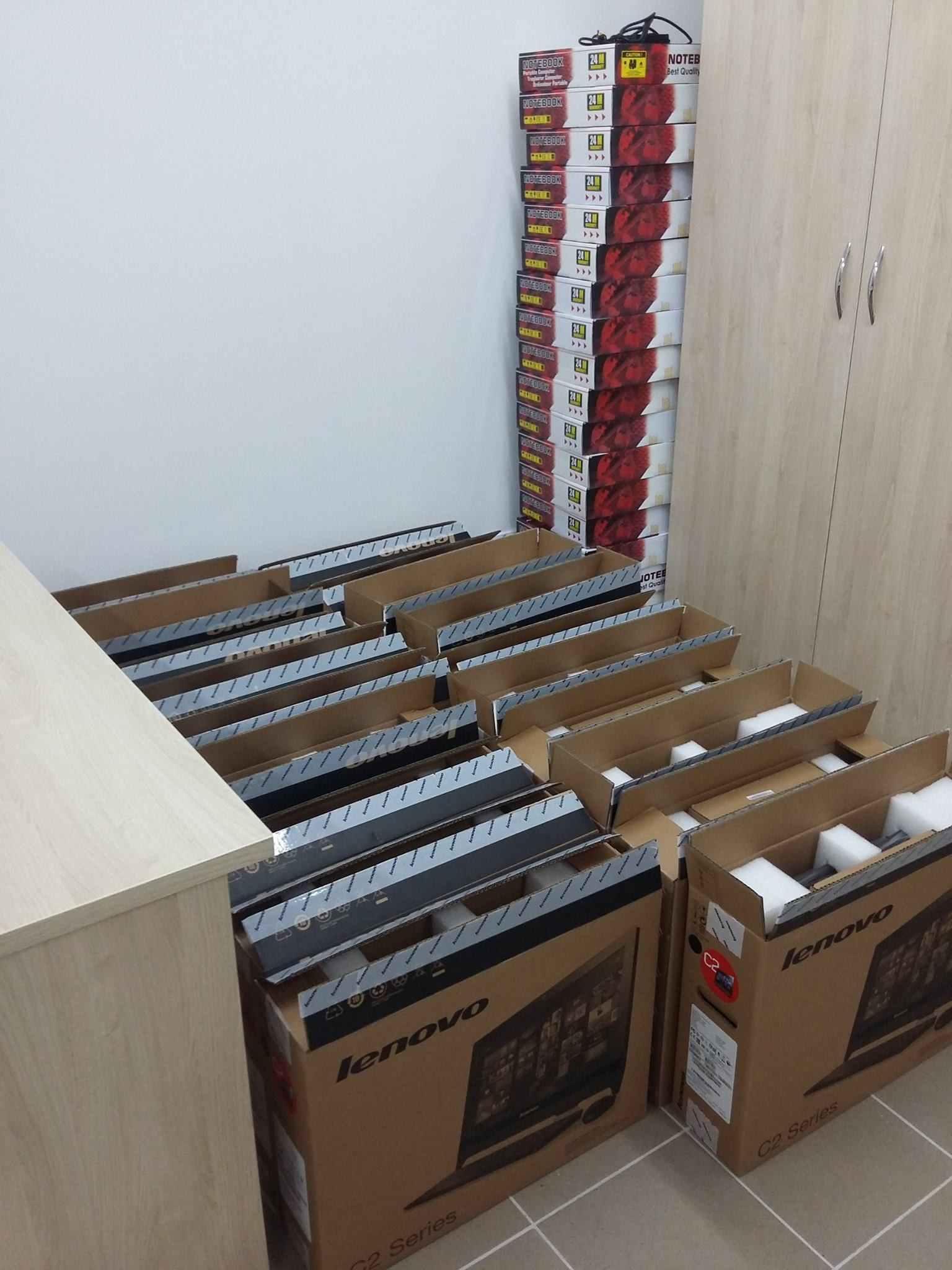 Stos kartonów z monitorami Lenovo C2 Series, częściowo rozpakowanych, z widocznym zabezpieczeniem piankowym, obok szafki i stosu pudełek z notebookami.