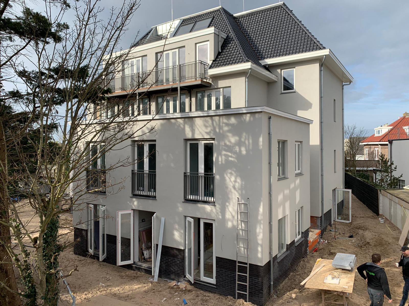 Nowoczesny, trzypiętrowy budynek z szarą elewacją, czarnym dachem i balkonami, częściowo wykończony, z otwartymi oknami na parterze i drabiną opartą o ścianę, widoczny z perspektywy placu budowy...