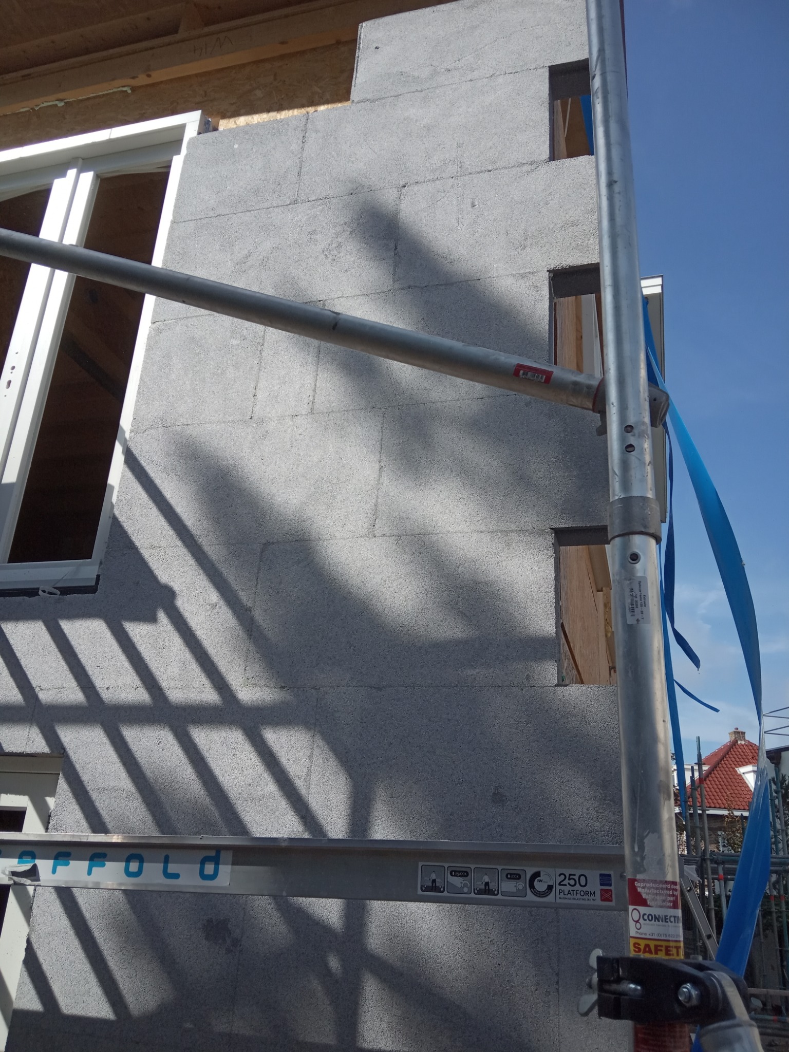 Ściana budynku w trakcie ocieplania, widoczne bloki izolacyjne, rusztowanie z niebieską taśmą zabezpieczającą, okno w stanie surowym.