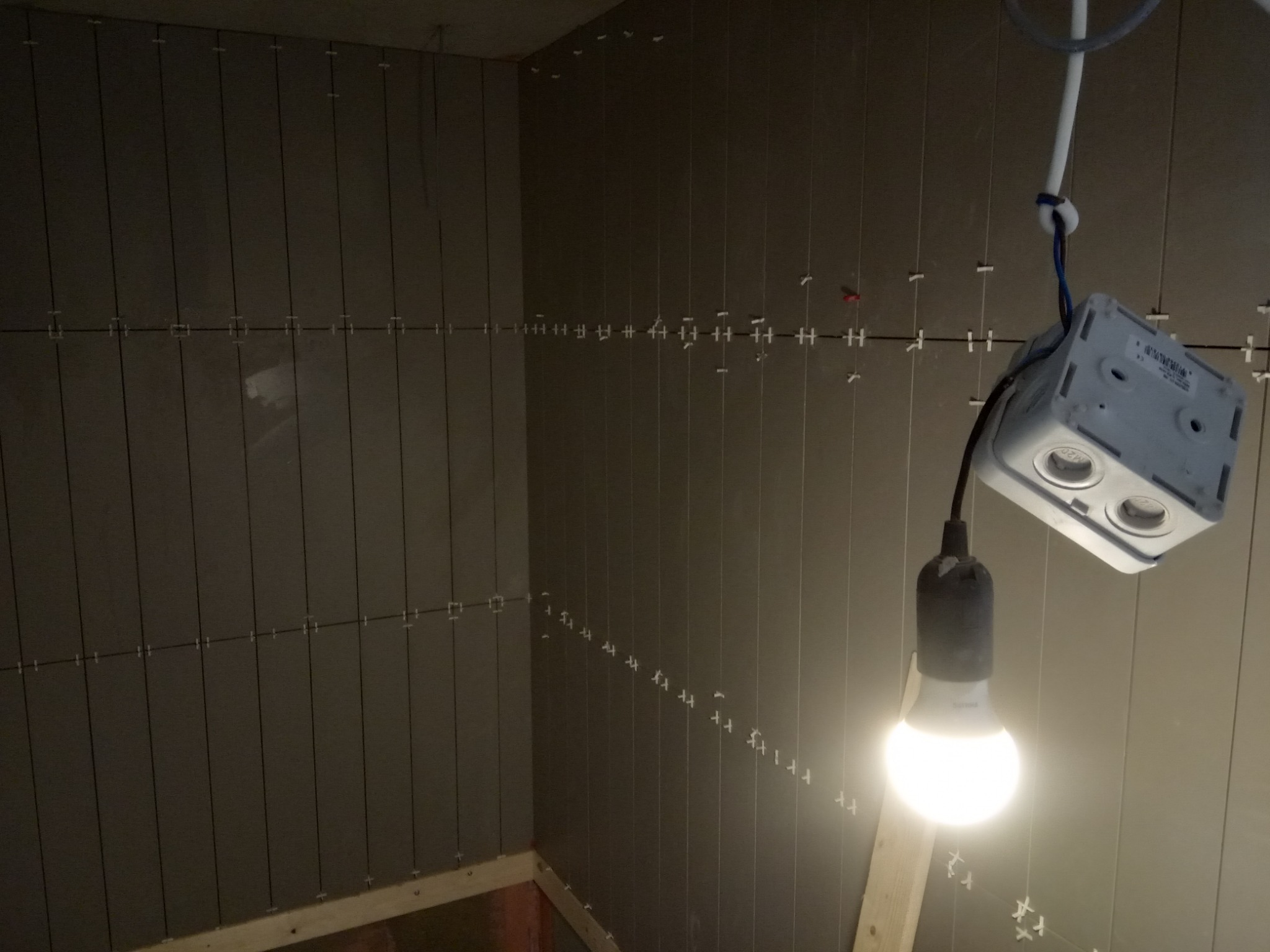 Świeżo ułożone, pionowe, ciemnoszare płytki na ścianie w rogu pomieszczenia, z widocznymi krzyżykami dystansowymi, oświetlone wiszącą żarówką.
