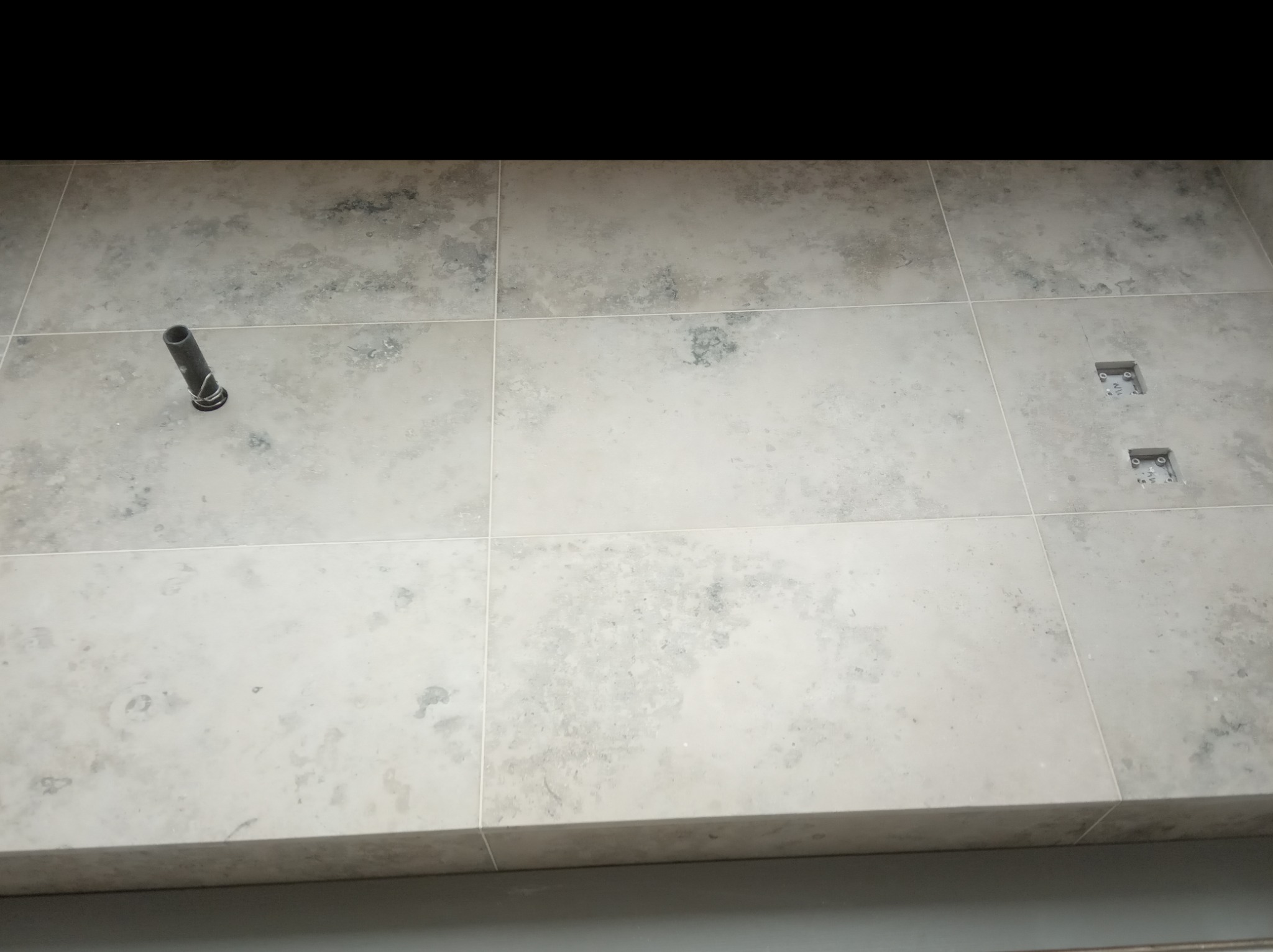 Ułożone jasne płytki podłogowe z widocznymi fugami, rurką przy ścianie i otworami na odpływ.