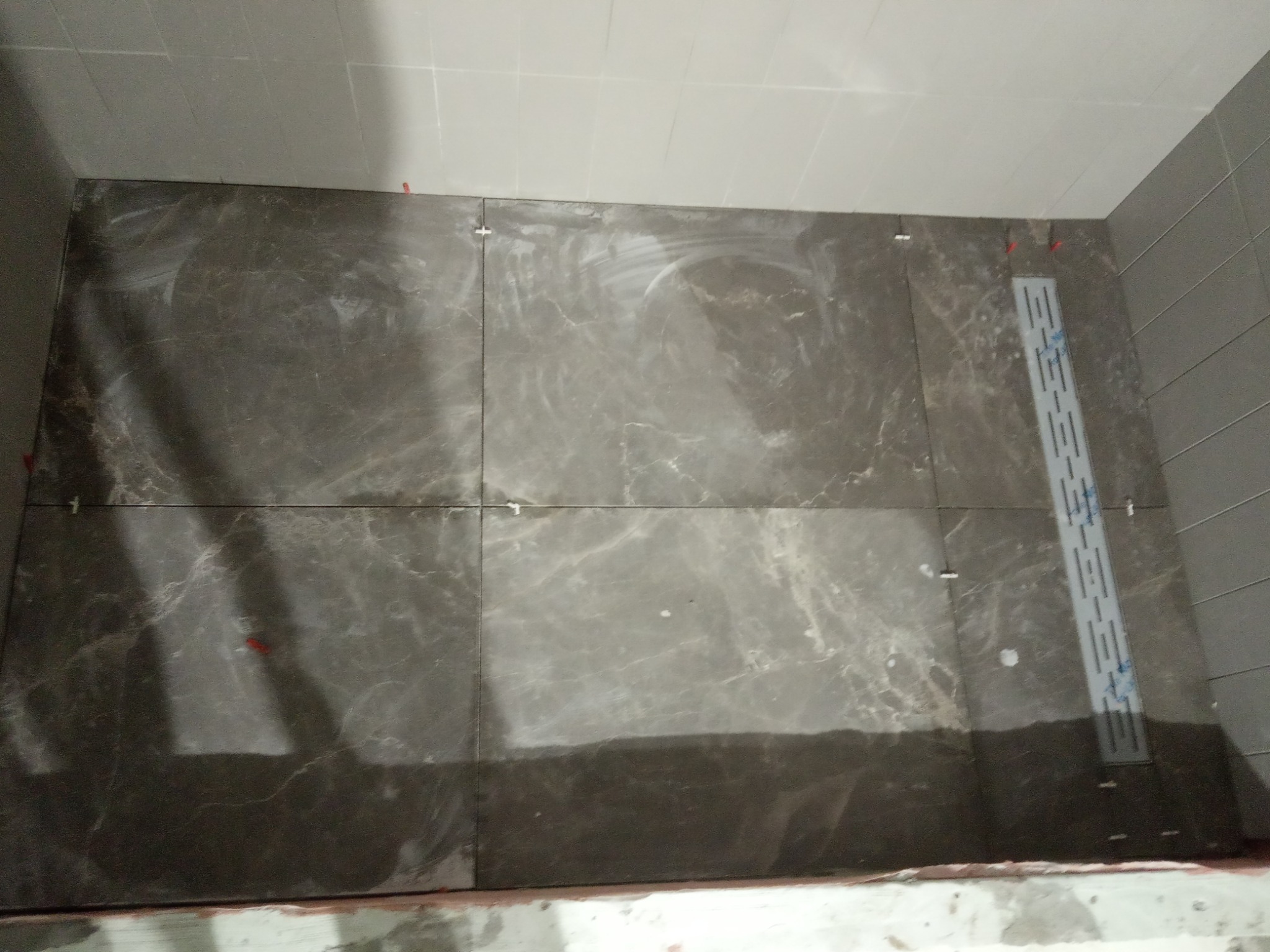 Układanie ciemnych, marmurkowych płytek w łazience, widoczny odpływ liniowy i krzyżyki dystansowe.