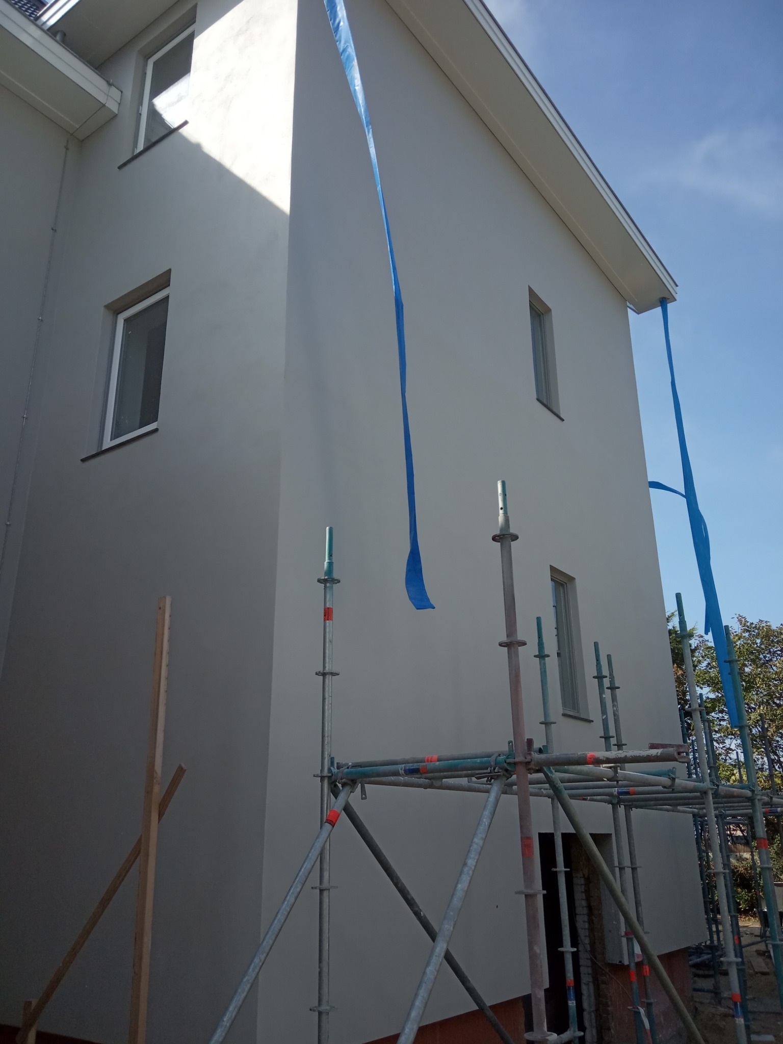 Świeżo pomalowana szara elewacja domu jednorodzinnego z widocznymi rusztowaniami i niebieską folią ochronną, w słoneczny dzień.