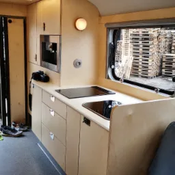 Jasna, drewniana zabudowa kuchenna w kamperze z wbudowanym ekspresem do kawy i płytą indukcyjną, widok na stosy palet przez okno.