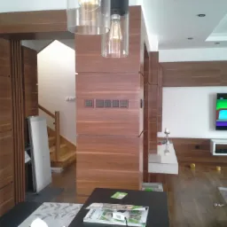 Nowoczesne wnętrze z elementami wykończenia z drewna orzechowego: zabudowa ściany z włącznikami, drzwi przesuwne i fragment schodów, oświetlenie punktowe i wiszące lampy, telewizor na ścianie...