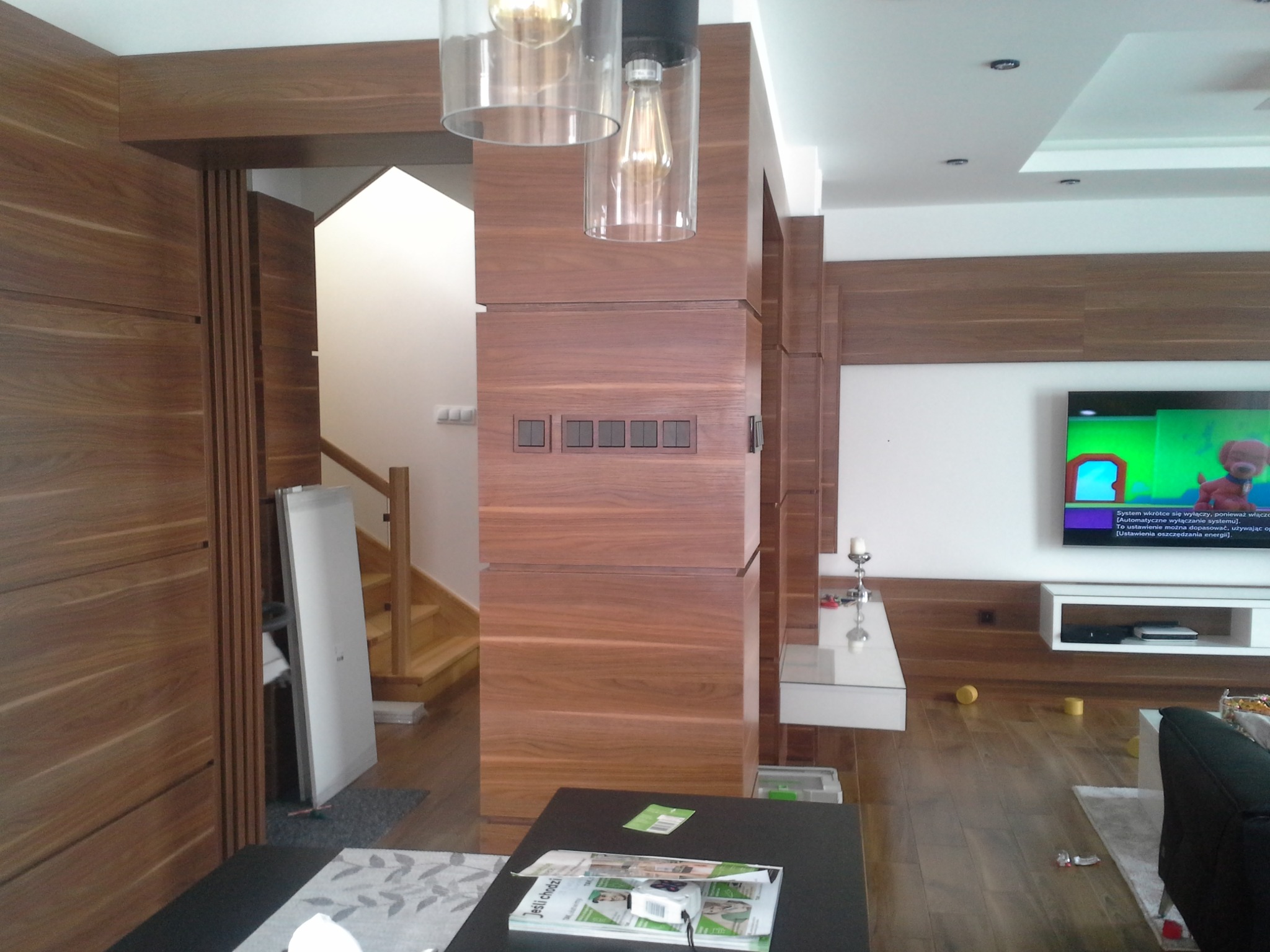 Nowoczesne wnętrze z elementami wykończenia z drewna orzechowego: zabudowa ściany z włącznikami, drzwi przesuwne i fragment schodów, oświetlenie punktowe i wiszące lampy, telewizor na ścianie...