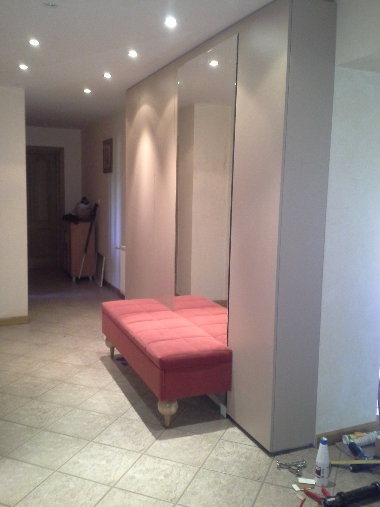 Jasnoszara szafa z lustrzanymi drzwiami i różową tapicerowaną ławką w przedpokoju; na podłodze widoczne narzędzia i akcesoria montażowe.