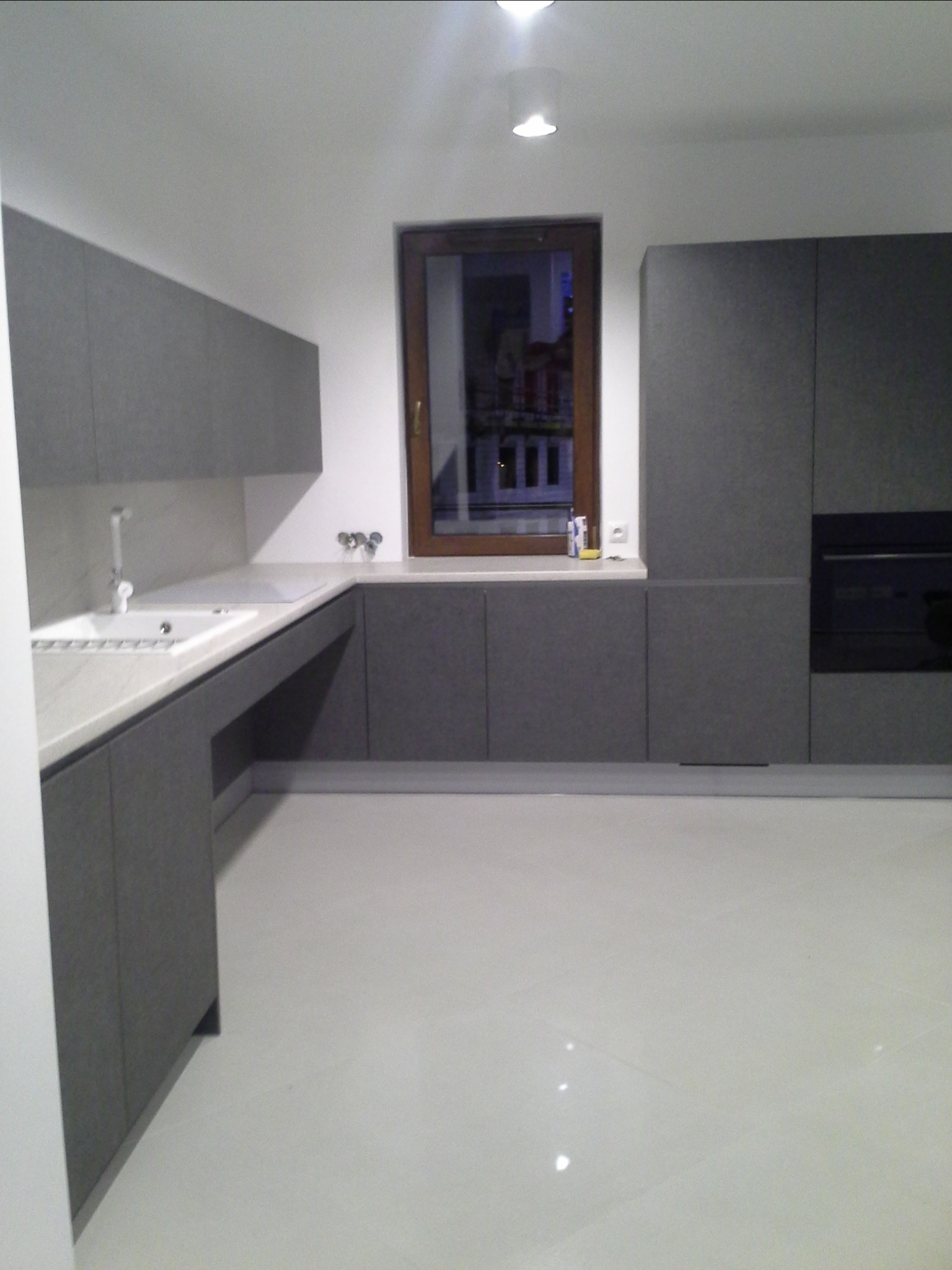 Nowoczesna kuchnia w odcieniach szarości z białym blatem i widokiem na nocne miasto za oknem.