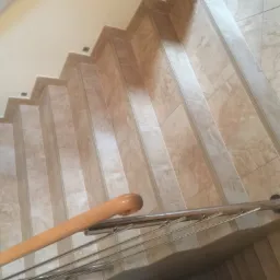 Wykończone schody wewnętrzne wyłożone beżowymi płytkami imitującymi marmur, z drewnianą poręczą i metalowymi wspornikami, oświetlone punktowymi lampkami w suficie.