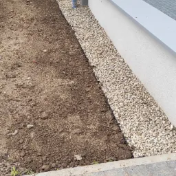 Przygotowany teren pod trawnik z wyłożonym obrzeżem z białego grysu, widoczna niebieska rura doprowadzająca wodę, kostka brukowa na pierwszym planie.