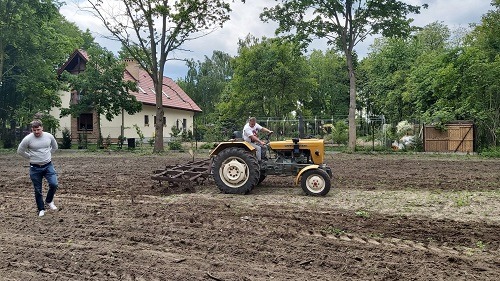 Żółty traktor z bronami talerzowymi na polu, przygotowujący grunt pod uprawę, obok stoi mężczyzna, w tle dom i zieleń.