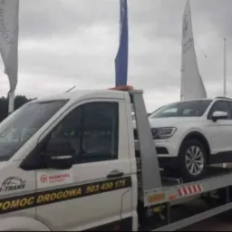Biała laweta z białym SUV-em Volkswagen Tiguan na platformie, widoczne logo firmy SPEED-TRANS POMOC DROGOWA i numer telefonu 503 430 175, w tle flagi reklamowe.