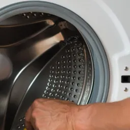 Technik w szarej odzieży roboczej z czerwoną lamówką przy kieszeni, pochylony nad otwartą pralką automatyczną podczas serwisu.