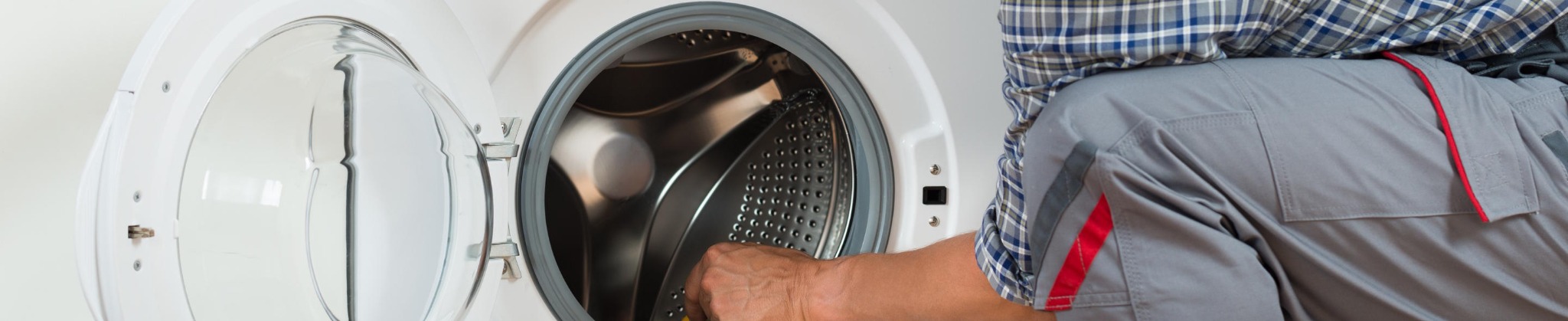 Technik w szarej odzieży roboczej z czerwoną lamówką przy kieszeni, pochylony nad otwartą pralką automatyczną podczas serwisu.