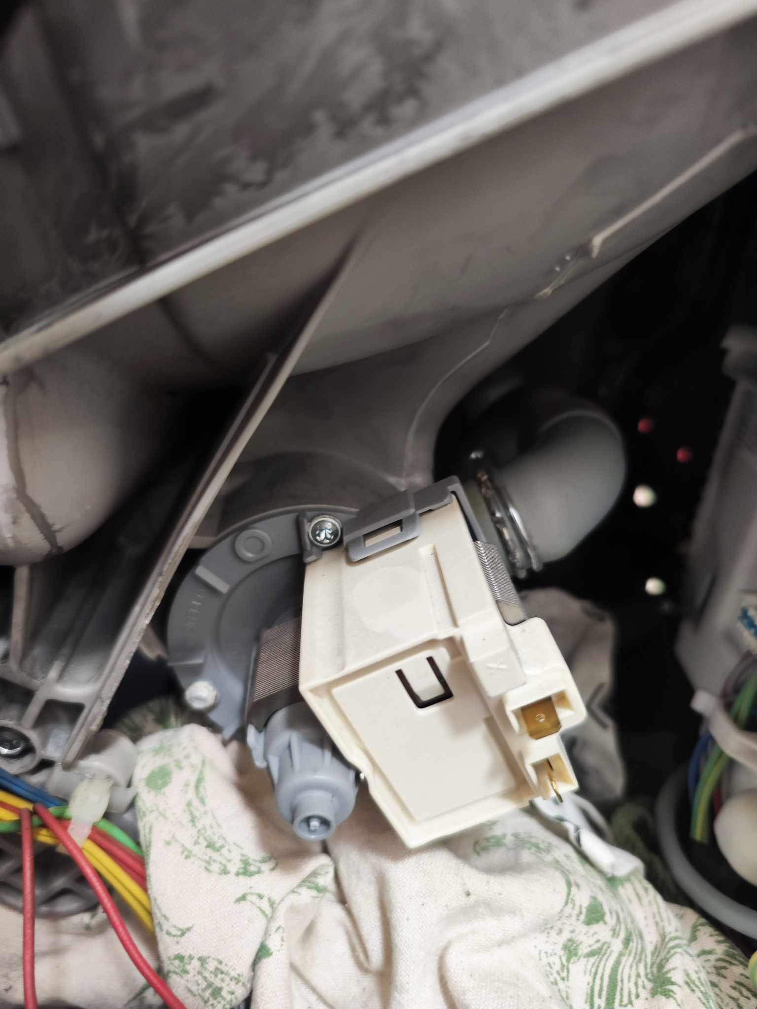 Wnętrze zmywarki w trakcie naprawy: widoczna pompa odpływowa z podłączonymi przewodami elektrycznymi oraz fragment węża odpływowego, całość osłonięta materiałem ochronnym.