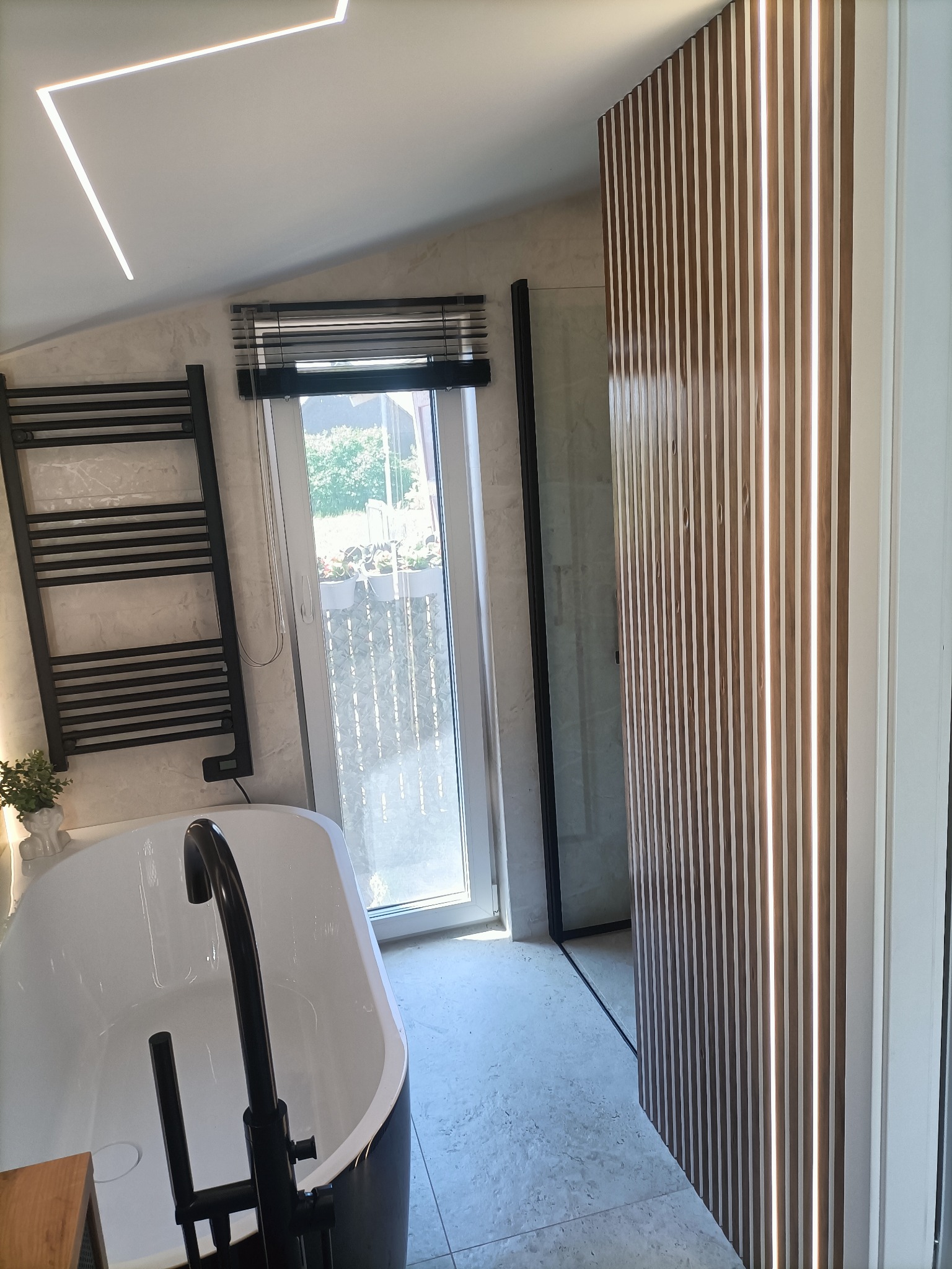 Nowoczesna łazienka z wolnostojącą białą wanną z czarną armaturą, grzejnikiem drabinkowym, drewnianą ścianką działową z podświetleniem LED oraz oknem z żaluzjami.