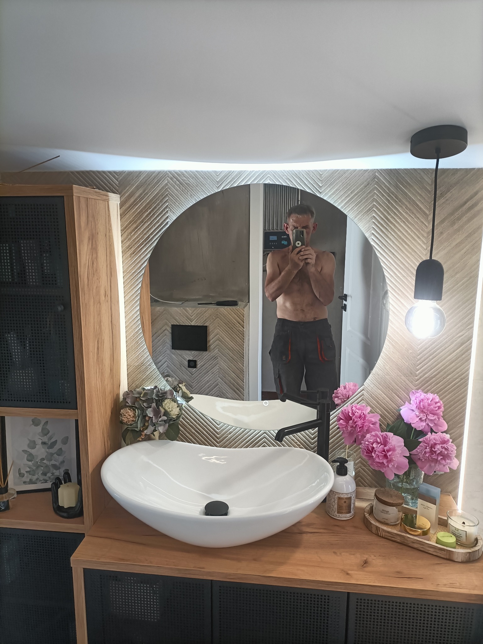 Nowoczesna łazienka z okrągłym lustrem podświetlanym LED, białą umywalką nablatową, drewnianym blatem, kwiatami i odbiciem fotografa w lustrze.