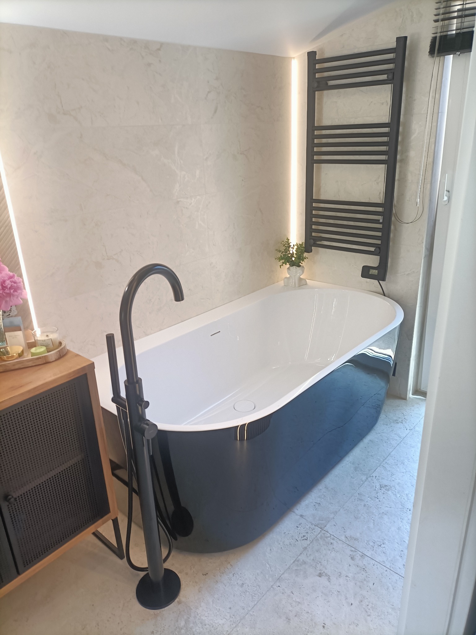 Nowoczesna łazienka z wolnostojącą czarno-białą wanną, czarną baterią podłogową i grzejnikiem drabinkowym, oświetlona ledowym paskiem i wykończona marmurowymi płytkami.