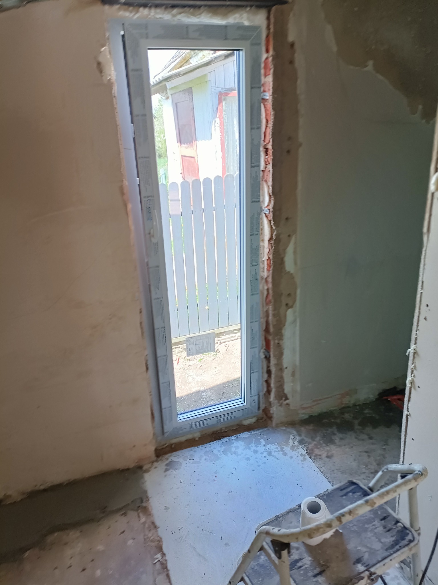Wnętrze w trakcie remontu łazienki: widok na nowo osadzone okno PCV z widokiem na biały płot, ściany w stanie surowym, drabina malarska z kubkiem.