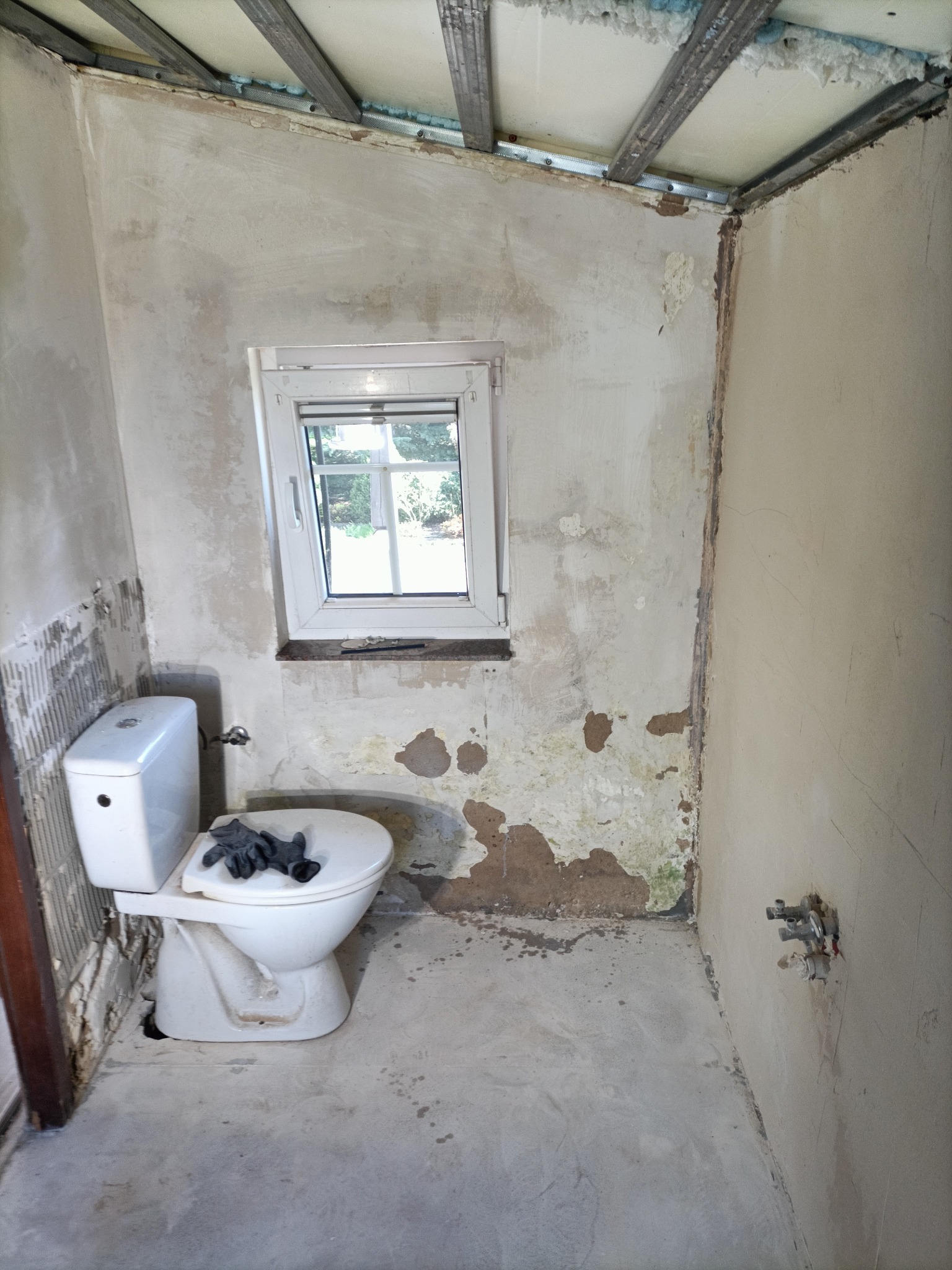 Łazienka w trakcie prac przygotowawczych: ściany z fragmentami starej glazury, surowy sufit podwieszany, biała toaleta z roboczymi rękawicami na desce, odsłonięte rury.