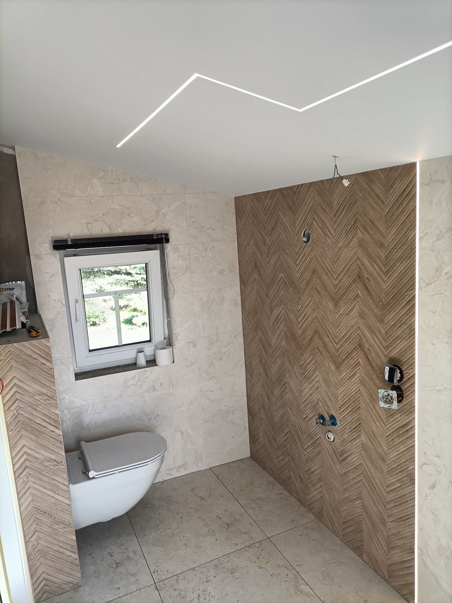 Wnętrze łazienki z toaletą pod oknem, ściany wyłożone płytkami imitującymi marmur i drewnem ułożonym w jodełkę, oświetlenie sufitowe LED.