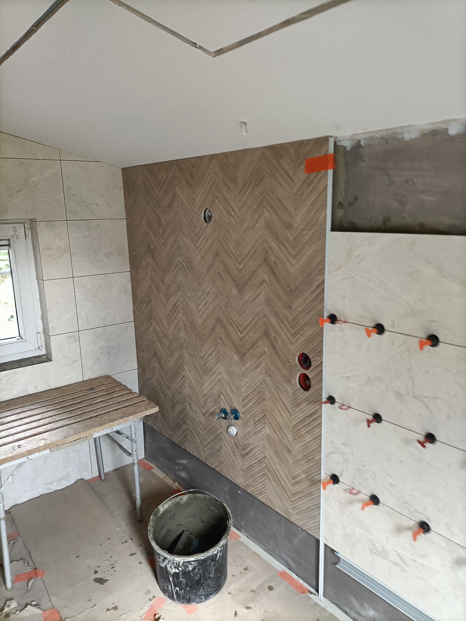 Ściana w łazience wyłożona płytkami w wzór jodełki, obok fragment ściany z płytkami mocowanymi za pomocą klipsów poziomujących, widoczne przygotowane instalacje wodne, drabinka i wiadro z zaprawą.