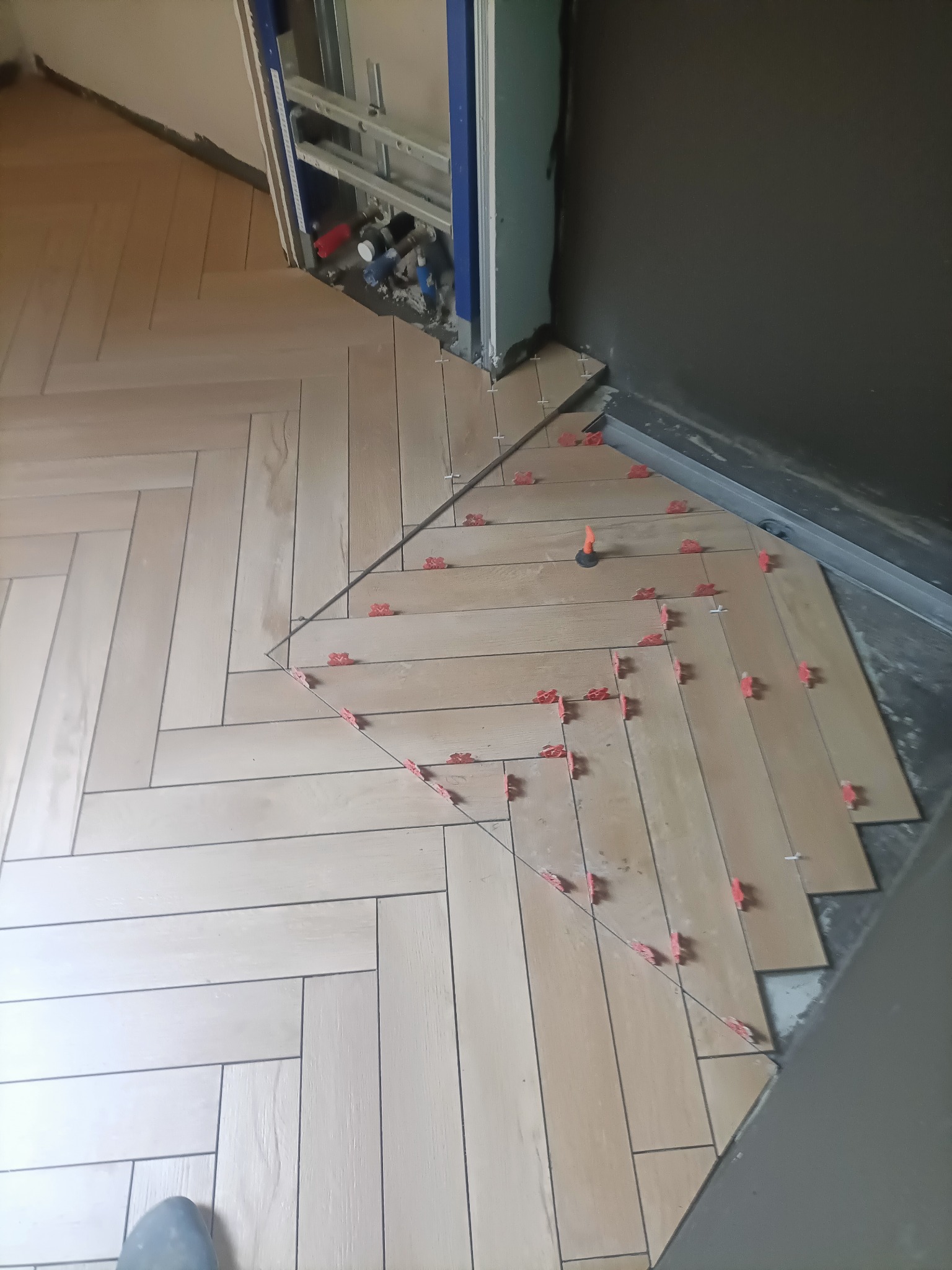 Układanie parkietu z jasnych płytek w jodełkę, z użyciem czerwonych krzyżyków dystansowych. Widoczne rury instalacyjne i fragment szarej ściany.