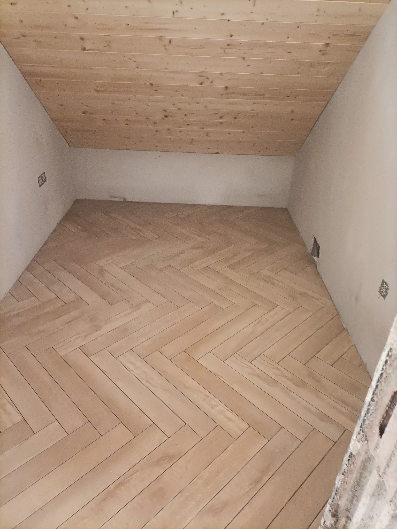 Jasna podłoga ułożona w jodełkę w pomieszczeniu z drewnianym sufitem. Widoczne gniazdka elektryczne i ściany w kolorze złamanej bieli.