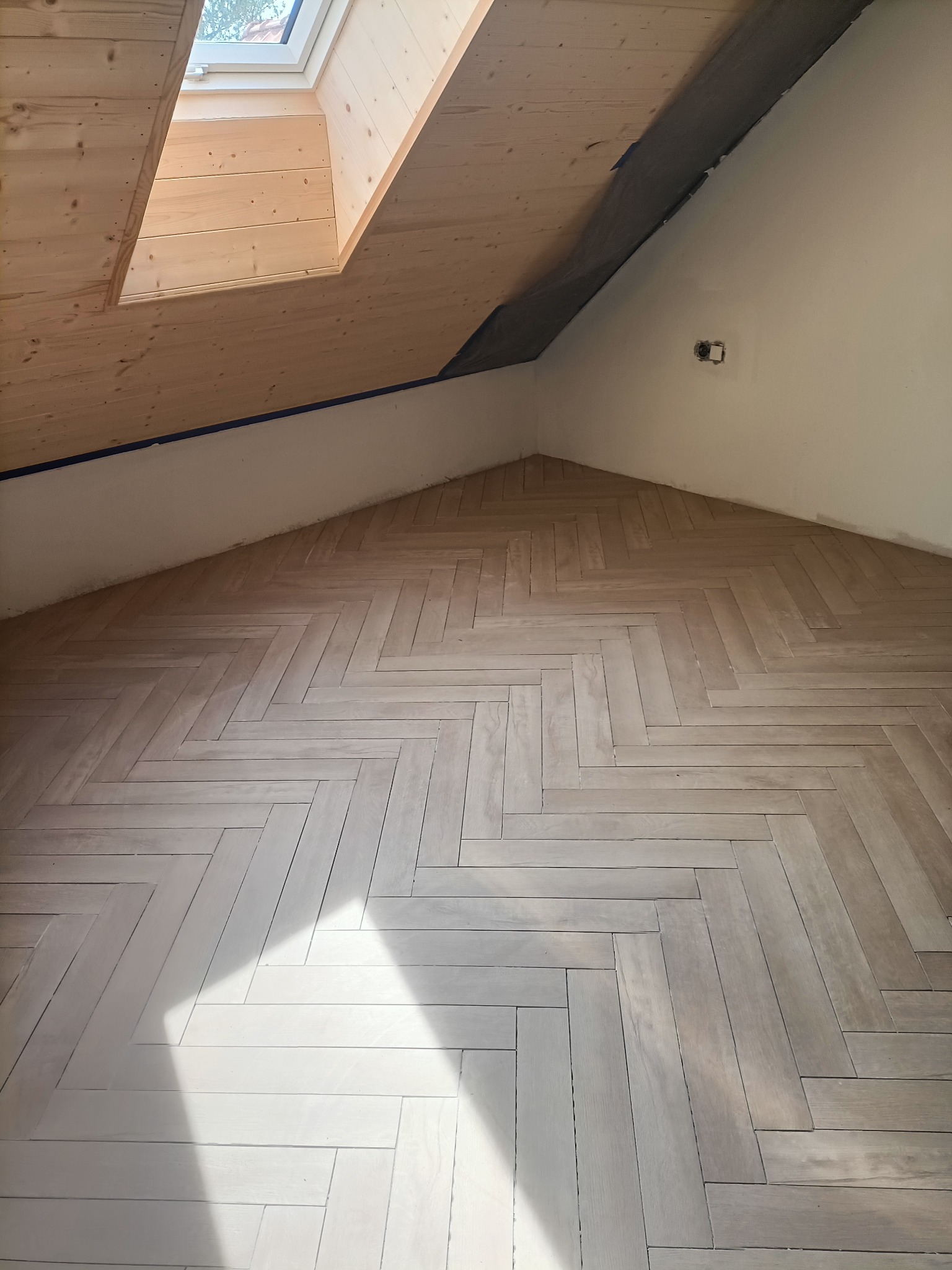 Jasna, drewniana podłoga ułożona w jodełkę w pokoju na poddaszu z oknem dachowym. Widoczne przygotowane ściany do malowania i drewniane wykończenie okna.