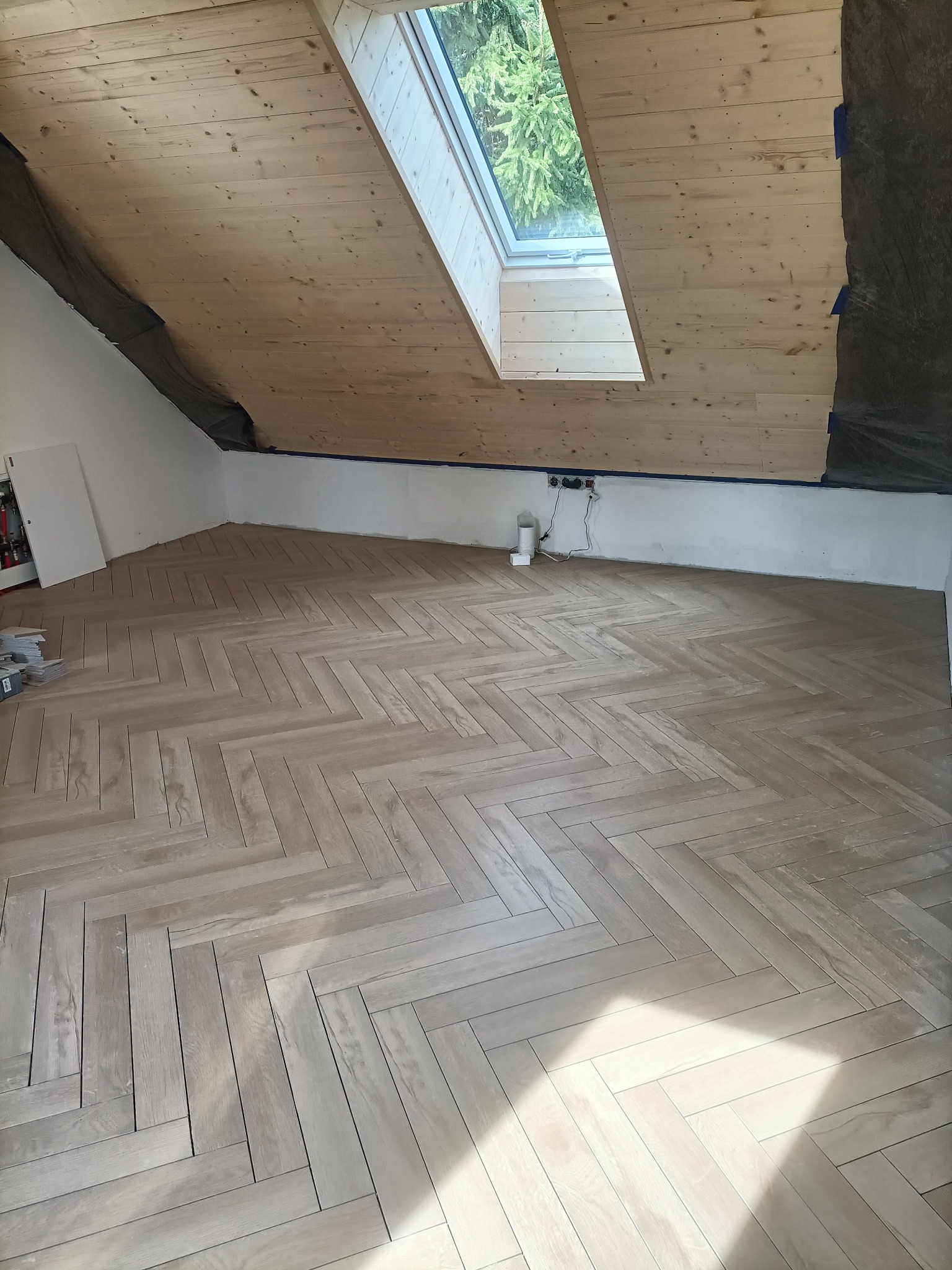 Jasne panele ułożone w jodełkę na poddaszu z oknem dachowym. Widoczne zabezpieczenia na ścianach i suficie. Gniazdka elektryczne w tle.