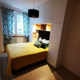 Nowoczesna sypialnia z zabudową meblową na wymiar: białe szafy z asymetrycznym bokiem, drewniane półki nad wezgłowiem łóżka, żółta narzuta i wzorzyste zasłony.