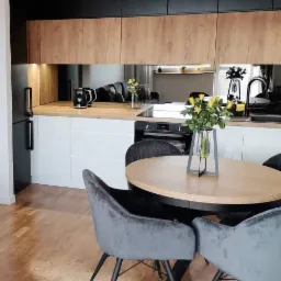 Nowoczesna kuchnia z drewnianymi frontami szafek wiszących, białymi szafkami dolnymi, czarną lod&oacute;wką i okrągłym stołem z szarymi krzesłami w tle odbijającym się w lustrzanym panelu.