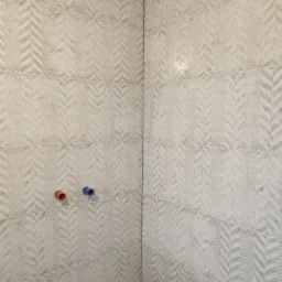 Wykończona ściana w łazience z beżowymi płytkami o wzorze jodełki, widoczne dwa wystające przyłącza wodne w kolorach czerwonym i niebieskim.