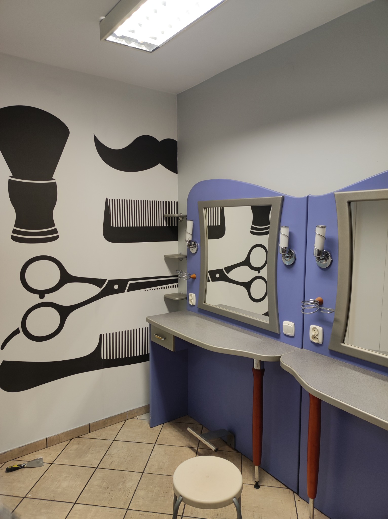 Wnętrze salonu fryzjerskiego z grafiką na ścianie przedstawiającą przybory fryzjerskie: pędzel, wąsy, grzebień i nożyczki, stanowiska z lustrami i oświetleniem, oraz taboretem.