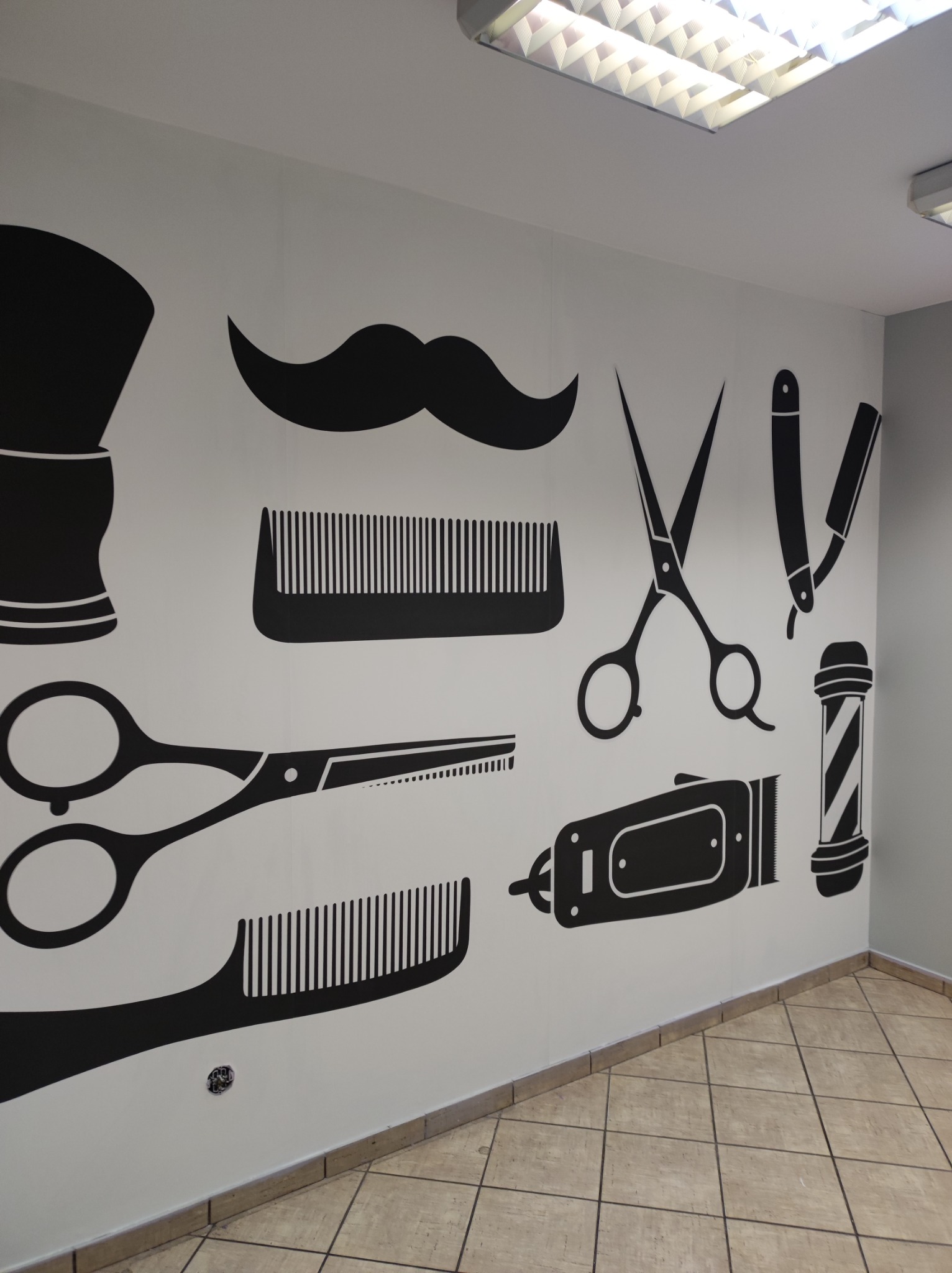 Ściana w salonie fryzjerskim ozdobiona czarnymi, graficznymi naklejkami przedstawiającymi akcesoria fryzjerskie: pędzel do golenia, wąsy, grzebień, nożyczki, brzytwę i maszynkę do strzyżenia.