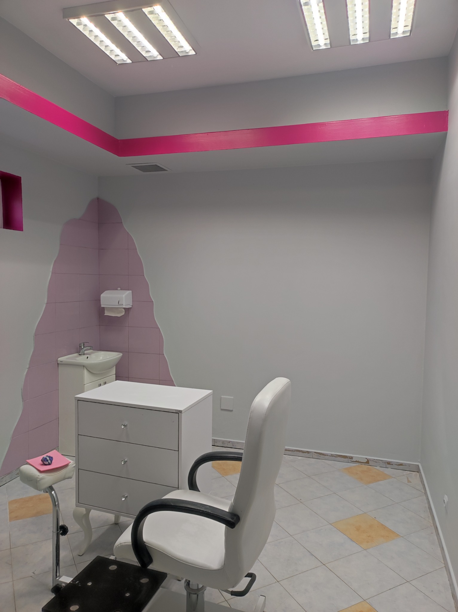 Wnętrze gabinetu kosmetycznego z fotelem, komodą z kryształowymi gałkami i umywalką w rogu, akcentowane różową listwą pod sufitem i dekoracyjną ścianą z różowymi płytkami.