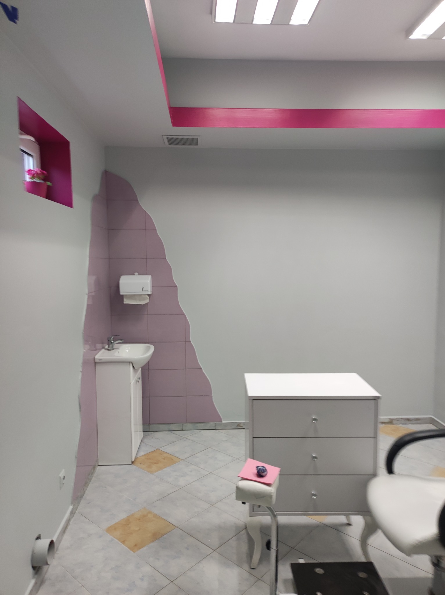 Wnętrze gabinetu kosmetycznego po odświeżeniu, z umywalką w narożniku wyłożonym fioletowymi płytkami, białą komodą z trzema szufladami, fioletowymi akcentami na suficie i ścianach oraz krzesłem...