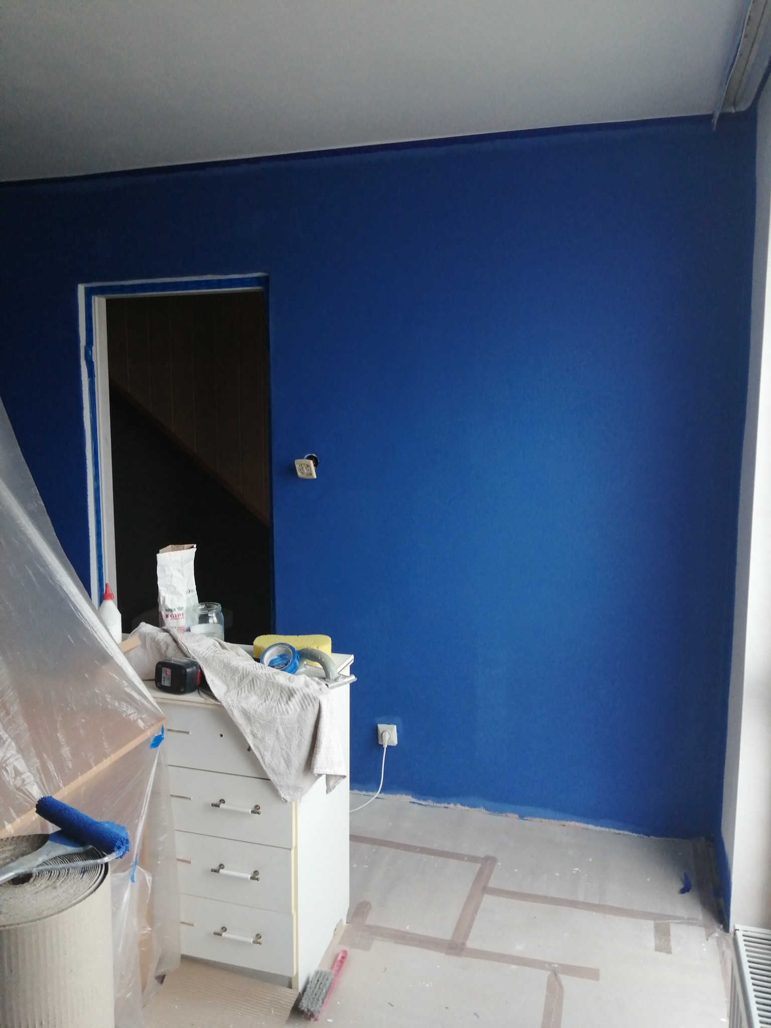 Pokój w trakcie odświeżania: ściana pomalowana na intensywny niebieski kolor, zabezpieczone folią meble i fragment podłogi, widoczne narzędzia malarskie i akcesoria na komodzie.