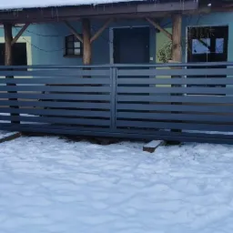 Nowoczesna, szara brama wjazdowa z poziomych profili, oparta tymczasowo na drewnianych klockach w zimowym krajobrazie.