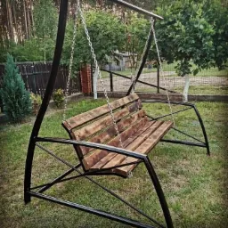 Metalowa huśtawka ogrodowa z drewnianym siedziskiem, łańcuchami i czarną ramą, umieszczona na trawniku w ogrodzie z widocznym ogrodzeniem panelowym w tle.