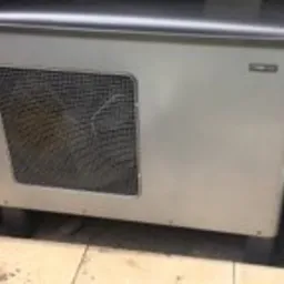 Biała jednostka centralna systemu wentylacji mechanicznej z odzyskiem ciepła, widoczna metalowa kratka wentylacyjna, szara obudowa, na podłodze widoczne fragmenty innych przedmiotów.