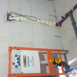 Stelaż podtynkowy WC marki JomoTech, zamontowany w surowym wnętrzu łazienki, widoczne rury instalacji wodnej i kanalizacyjnej, ściana z bloczków z naniesionymi odręcznymi notatkami technicznymi.